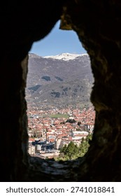 Through an ancient arrow slit window of Samuel's Fortress, Ohrid's red-roofed houses and distant snow-capped mountains are perfectly framed on a bright day.