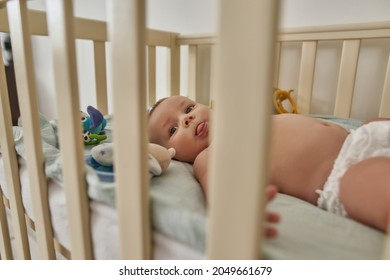 Through Crib Bars View Funny Infant Baby, Lying On Back And Showing Its Tongue. Baby Licking Lips Showing First Feeding Signs.