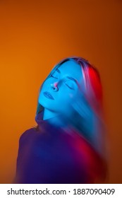 Through Glass Of Calm Young Female Model With Long Straight Hair Looking At Camera Against Orange Background In Studio With Blue Neon Light