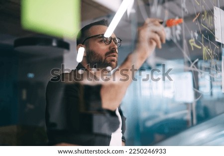 Through glass view of concentrated bearded male employee in eyeglasses pointing at data with marker and explaining project details to audience in meeting room in office