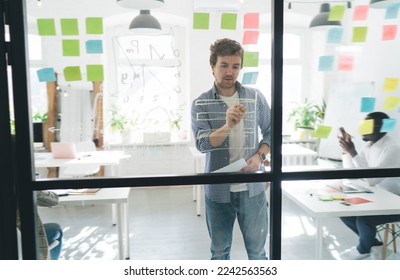 Through glass view of positive young male employee while working with African American and faceless coworker standing near glass wall with sticky notes and writing details in office - Powered by Shutterstock