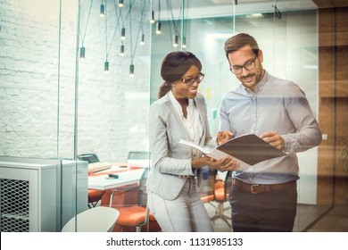 Through glass view of two smiling multi-ethnic business colleagues reviewing paper documents while standing in modern office - Powered by Shutterstock