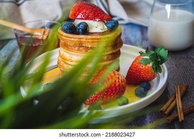 Through The Grass View Of Stack Of Pancakes With Honey Stick, Strawberry, Blue Berry And Whip Cream On Top In Plate, Delicious Dessert With Milk For Breakfast.