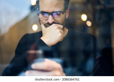 Through window of crop ethnic bearded thoughtful male in casual outfit and glasses sitting in modern cafe and surfing smartphone - Powered by Shutterstock