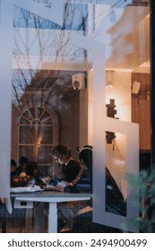 Through the window, a glimpse into a warm, dimly-lit office space with focused individuals working in the evening creates a serene work environment.