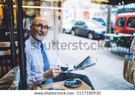 Through window senior short haired male in white shirt with coffee cup and newspaper sitting and looking away on city street near road