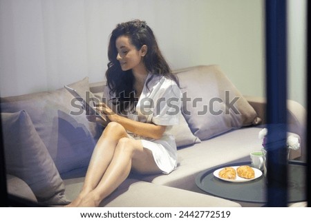 Through the window shot. Caucasian cheerful young woman with long hair sitting on couch in modern room typing on tablet device. Happy attractive female in white robe using gadget Stock fotó © 