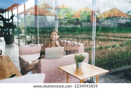 Through window of thoughtful female freelancer with long hair sitting on couch and leaning on hand while working on remote project using laptop in cafe