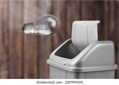 Throw A Light Bulb In A Trash Can, A Gray Bucket On A Wooden Background With A Light Bulb Thrown Into It, A Bad Idea Concept	