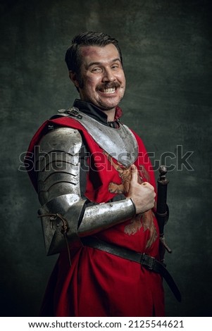 Thumb up. Portrait of smiling man in image of medieval warrior with wounded face wearing equipment isolated over dark background. Comparison of eras, history, renaissance style. Funny meme emotions