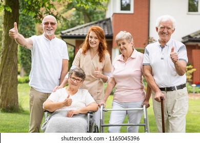 Thumbs up shown by happy female and male seniors and a caregiver outside their nursing home.