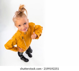 Thumbs Up. Pretty Blonde Little Girl In A Leather Mustard Jacket, Black Snake Texture Leggings And Boots. High Fashion, Full Length Standing Pose, Isolated Against A Studio Background