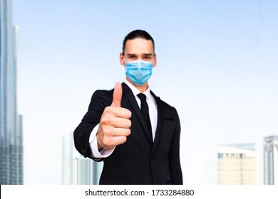 Thumbs Up! Young Male Businessman Giving Approval Hand Gesture To The Camera And Wearing Blue Anti Virus Medical Mask. Business Man Portrait With Modern City Blue Sky In Background.