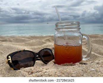 Thunder Bay, Barbados - May 25, 2022: A Glass Mason Jar With A Straw Filled With Rum Punch And Sunglasses On A Beach With Dramatic Clouds In The Background