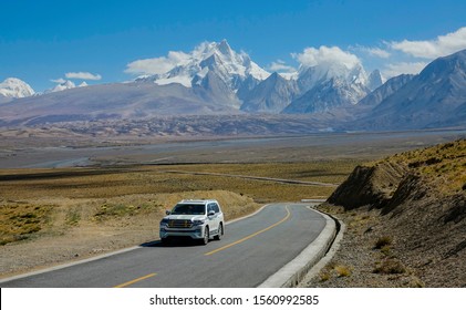 TIBETAN PLATEAU, CHINA, OCTOBER 2018: Local people driving across the breathtaking plains of the Himalayan Plateau. White SUV drives down an empty road and away from the snowy mountains in Tibet.