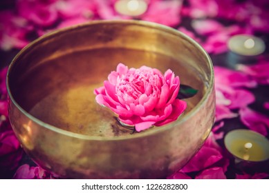 Tibetan Singing Bowl With Floating In Water Lily Inside. Special Sticks, Burning Candles, Lily Flowers And Petals On The Black Wooden Background. Meditation And Relax. Exotic Massage. Selective Focus