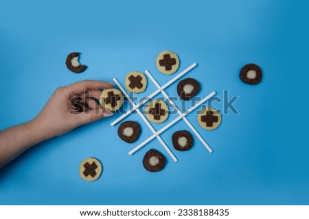 Tic tac toe game with freshly baked cookies and a woman's hand reaches for one of the cookies on blue background top view
