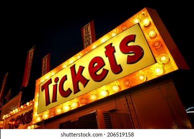 Tickets advertising neon sign with bright yellow lights marquee above carnival ticket counter booth and distributor vending machine at a festive county fair at night