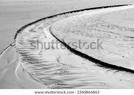 Tidal creek at low tide on the sandy beach of Juist island. Water flowing towards the North Sea and current forming a bent temporary river bed. Small natural structures and waves, black and white. 