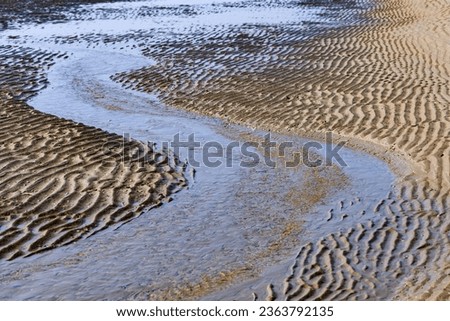 Tidal creek at low tide on the sandy beach of Juist island. Water flowing towards the North Sea and current forming a meandering s-curved temporary river bed. Small natural structures and waves.