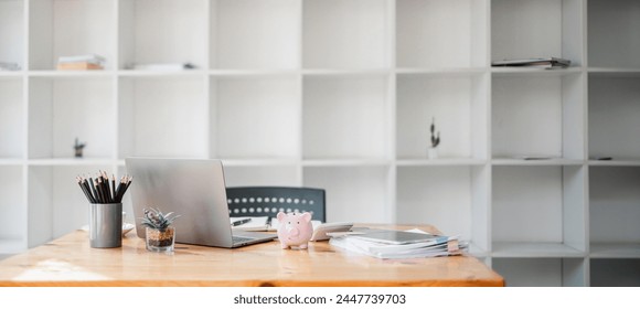 Tidy workspace featuring a laptop, colorful pencils in a holder, and a piggy bank, set against a backdrop of white shelves. - Powered by Shutterstock