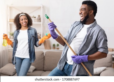 Tidying With Fun. Joyful Black Couple Fooling While Cleaning Flat Together, Man Using Mop Handle As Guitar And Laughing