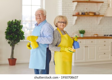 Tidying Up Your Home. Happy Senior Couple In Yellow Aprons And Gloves Smiling And Holding Detergent Bottles Standing Back To Back In The Kitchen After Cleaning Their House