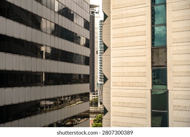 A tight architectural frame capturing the contrast between two close-proximity high-rise buildings in a metropolis. On the left, a modern commercial building features dark tinted glass windows that reflect the surrounding urban environment. On the right, a textured beige concrete facade shows off structural patterns and vertical glass paneling. Other towers peek through the narrow gap between them, illustrating a dense urban landscape. Perfect for real estate, corporate, and city planning concepts.