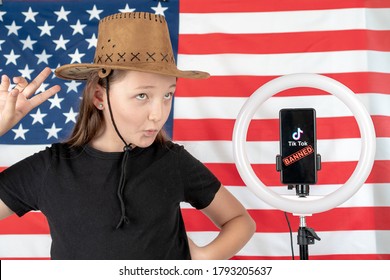 Tiktok Logo On A Smartphone. A Girl In A Cowboy Hat Is Dancing Nearby And Taking A Selfie Against The Background Of The American Flag.  Russia, Kazan - August 08, 2020.
