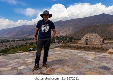 Tilcara, Jujuy, Argentina. 02-03-2022. Adult Male (45-50 Years Old) In Hiking Clothes And Hat On Top Of A Mountain, With Another Colorful Mountains In The Background.