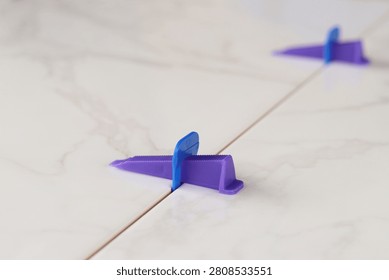 Tile leveling process, tile leveling system, wedge close-up. Purple plastic wedge and blue clip are installed between two marble-textured tiles. Modern tools for floor tiling