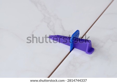 Tile leveling process, tile leveling system, wedge close-up. Purple plastic wedge and blue clip are installed between two marble-textured tiles. Modern tools for floor tiling
