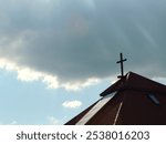 The tiled roof of a Protestant church with a cross on top against a blue sky with a large gray cloud in the background