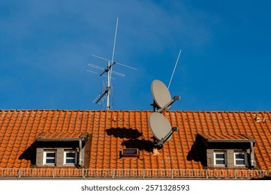 A tiled rooftop features two large satellite dishes alongside an antenna against a bright blue sky.