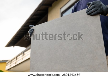  tiling work. Man carrying large paving tile for patio installation near house. Construction worker transporting a large tile slab for terrace . professional tiling work.