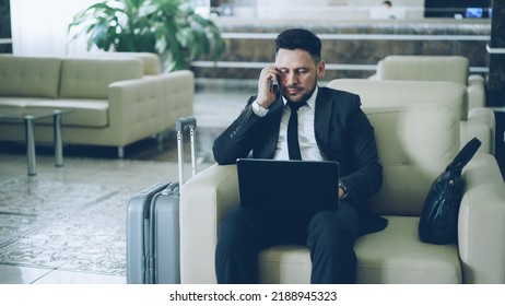 Tilt up of confident attractive businessman talking mobile phone and using laptop computer while sitting on armchair in luxury hotel with luggage suitcase near him. Travel, business and people
