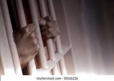 Tilt view of a black man's hands gripping the bars of a prison cell. Horizontal format.