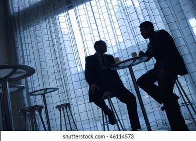 Tilt View Silhouette Of African-American And Asian Businessmen Sitting At A Table Having Coffee In Front Of A Curtained Window.