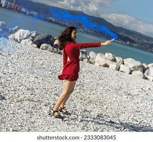 tilted shot of a slim girl in a red dress holding a blue smoke flare near the sea - Powered by Shutterstock