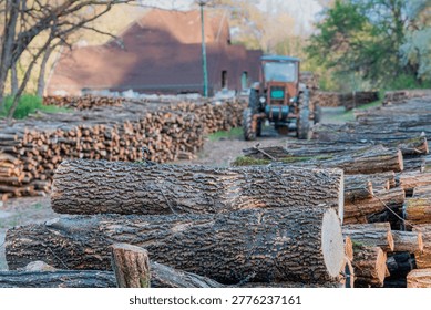 Timber logs waiting for processing, stacked in foreground with an agricultural tractor visible in the blurred background
