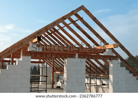 A timber roof truss in a house under construction, walls made of autoclaved aerated concrete blocks, a rough window opening, a reinforced brick lintel, a scaffolding, blue sky in the background