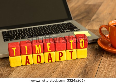 Time To Adapt written on a wooden cube in front of a laptop
