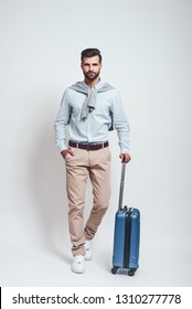 Time For Adventure. Full Length Of Charming Bearded Young Man With A Luggage Keeping One Hand In His Pocket And Looking At Camera. Travel Concept. Grey Background