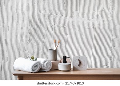 Time for beauty. Concepts of morning routine at home. Bathroom table with white alarm clock, rolled fresh towels, soap dispenser and bamboo toothbrush in ceramic cup against textured copy space wall