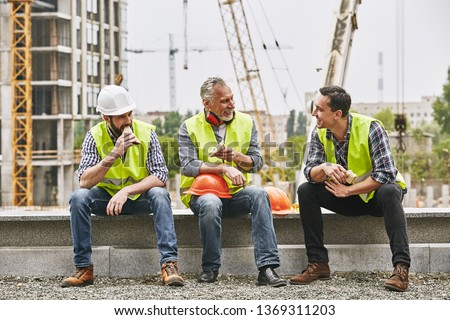 Time for a break. Group of builders in working uniform are eating sandwiches and talking while sitting on stone surface against construction site. Building concept. Lunch concept