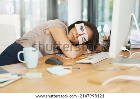 Time for a catnap. Shot of a tired businesswoman napping at her desk with adhesive notes on her eyes.