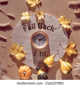 Time change in Autumn. Text Fall Back 1 hour on circle stone board. Wooden alarm clock, with dry Fall leaves on recycled cardboard. Top view, flat lay, sunlight with long shadows.