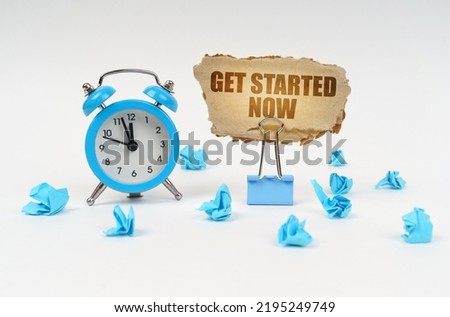 Time concept. On a white surface, an alarm clock, an office clip, crumpled paper and a cardboard sign with the text - Get Started Now