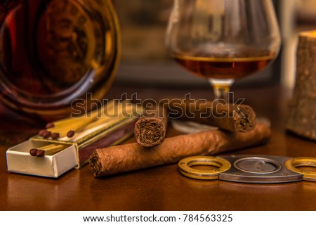 Time to enjoy! Quality cigars and cognac on a wooden table and other accessories such as cigar cutters and matches. In the background a fireplace. Focus on the cigars. Concept: life style or health
