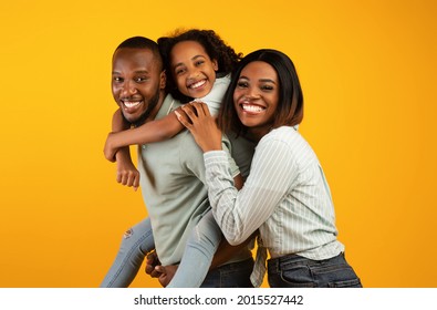 Time with family. Overjoyed african american family laughing and posing to camera isolated over yellow studio wall. Cheerful father carrying his daughter on back